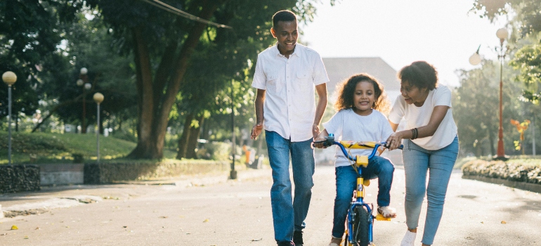 A smiling family plays in a park with a kid on the bike.
