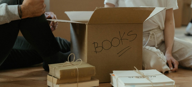 A man and a woman are sitting on the floor and packing books into a carton box.